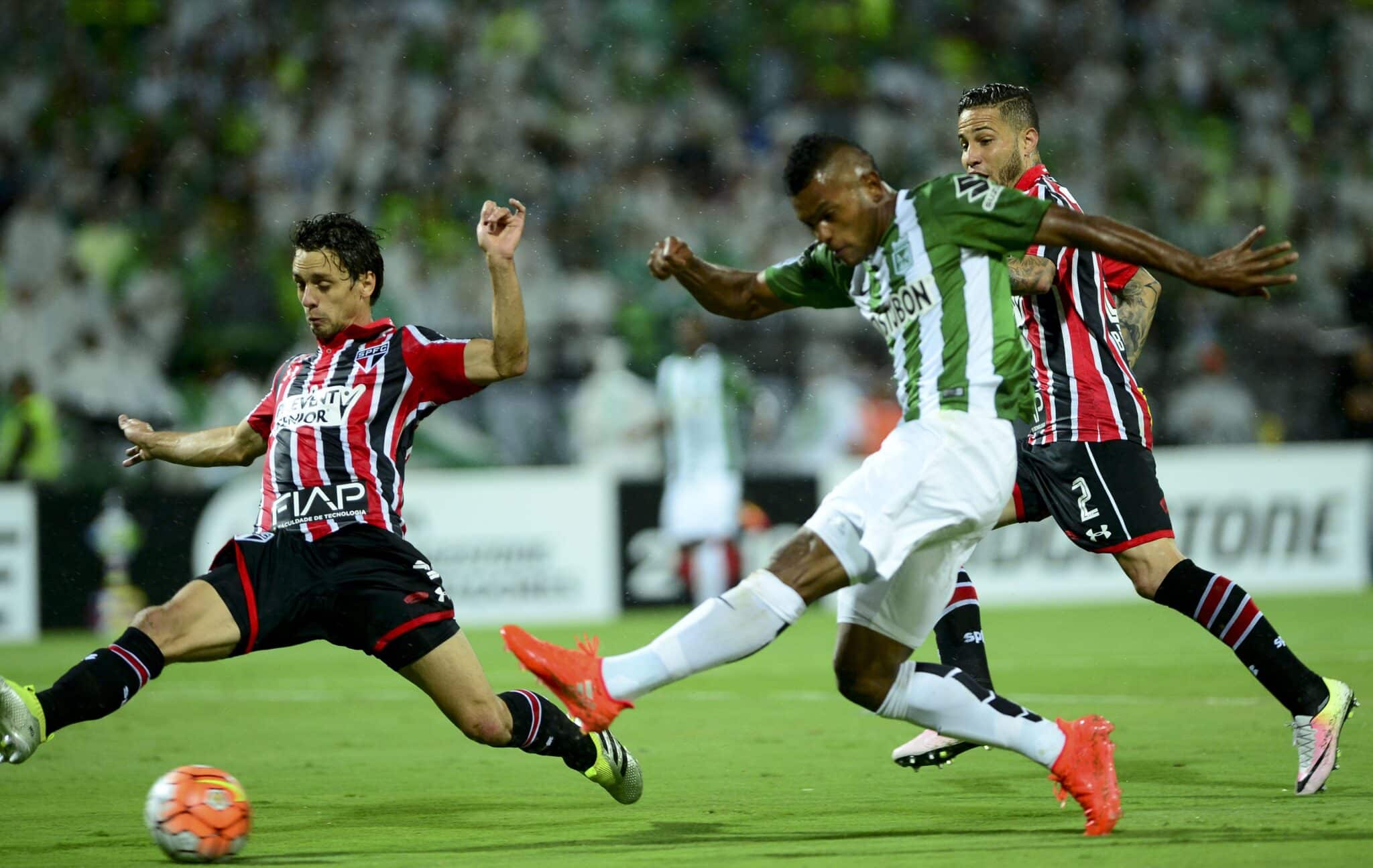 Jogador do Atlético Nacional chuta a bola marcado por zagueiro do São Paulo